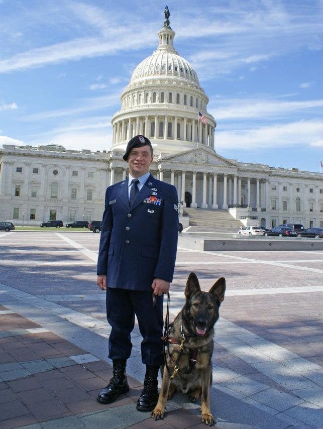 SrA Michael M. in uniform and sable guide dog "Xxon" mouth open tongue out standing in front of the US capitol building.