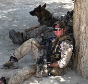 Brad S in military fatigues sitting on ground with comrade and German Shepherd sitting next to him