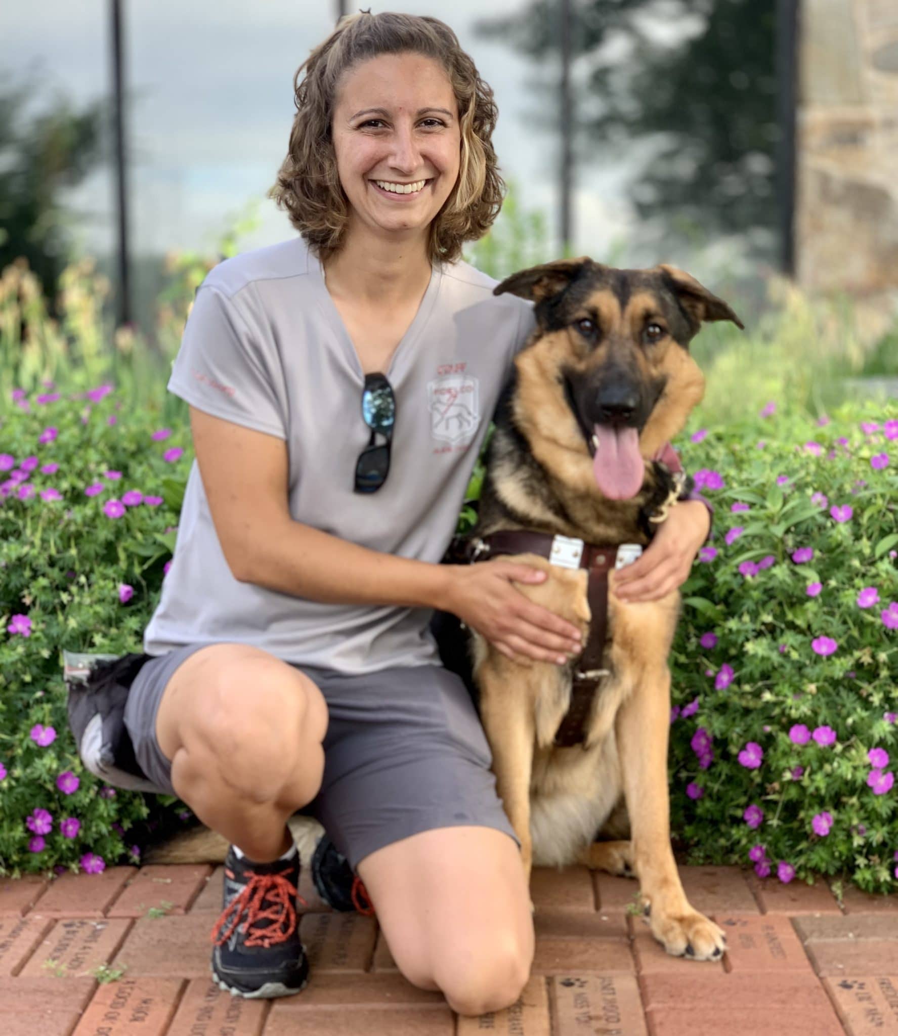 Trainer Chelsea, in gray Fidelco shirt and shorts smiling and kneeling next to guide dog in training "Bella" in harness and tongue out