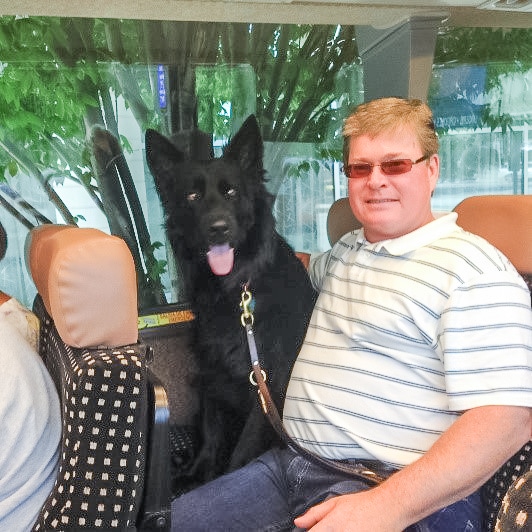 Retired US Air force master sgt gary traynor sits (on right) on a bus smiling at the camera next to his black GSD guide dog Reuben who has is mouth open and tongue out. 