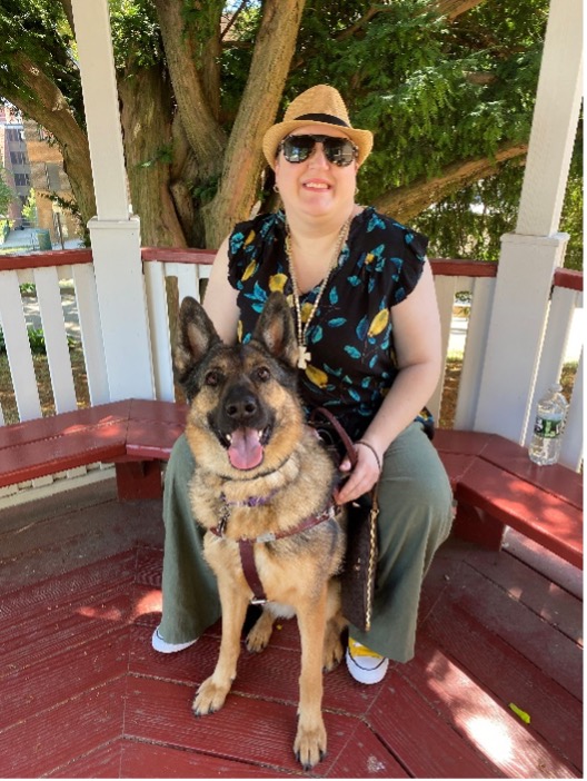 Holly smiles in sunglasses in a hat sitting with Teagan in harness looking up at camera