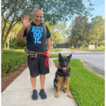 Yehia waving holding the leash of guide dog vader on the sidewalk