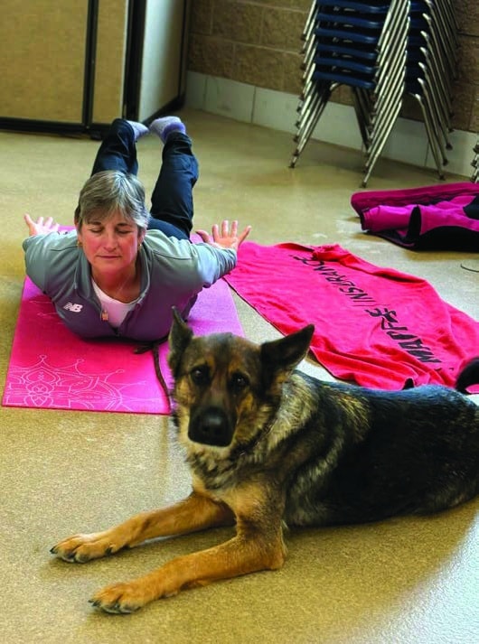 Kim doing yoga pose on stomach with her eyes closed while "Tori," a sable GSD, laying in front of her, looking at camera.