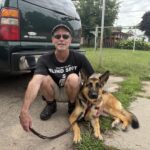 Bryan crouched down with koda his guide dog in harness laying on the side walk