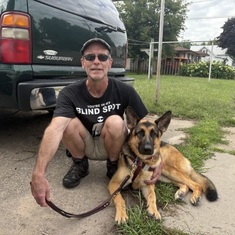 Bryan crouched down with koda his guide dog in harness laying on the side walk