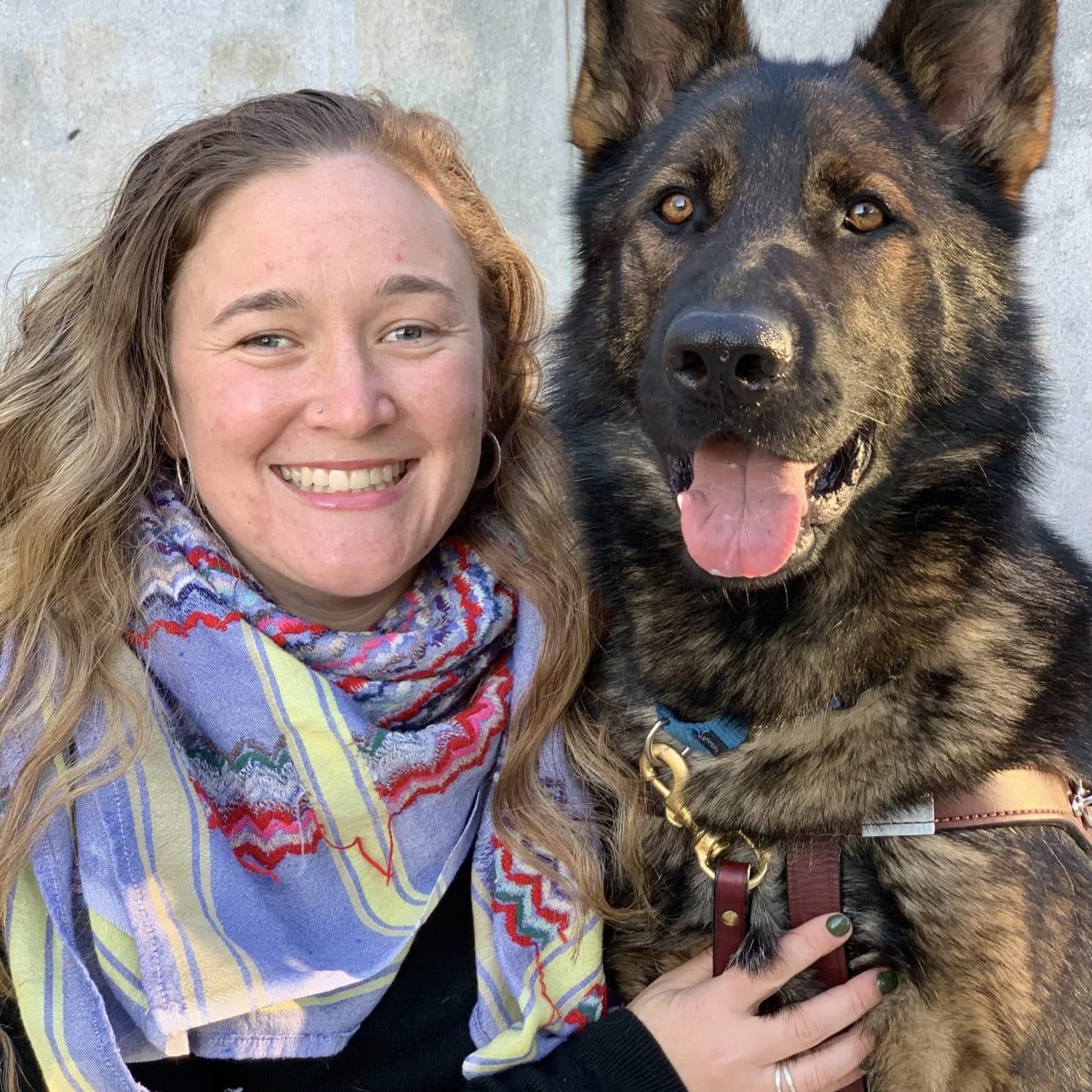 Maggie 32 year old female blonde women smiles with one hand on Gus' chest, her dark sable guide dog in harness looking at the camera